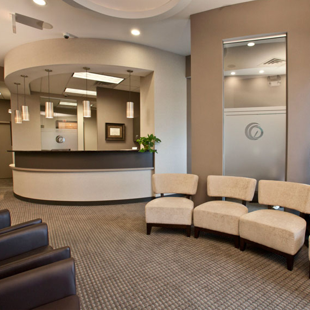 The image shows an interior space that appears to be a waiting area or reception within a professional office environment. There is a reception desk, a seating area with multiple chairs and ottomans, a wall-mounted television screen, and decorative elements such as plants and artwork.